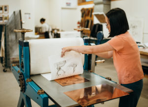 A woman pulling a print from a copper etching plater at a press.