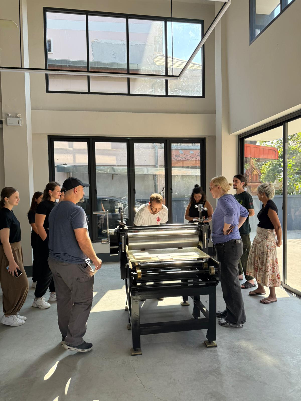PHOTO-2025-06-14-02-30-30 3 A group of people gathered around the etching press at Hello Print Friend Studio surrounded by large windows letting in lots of natural light.