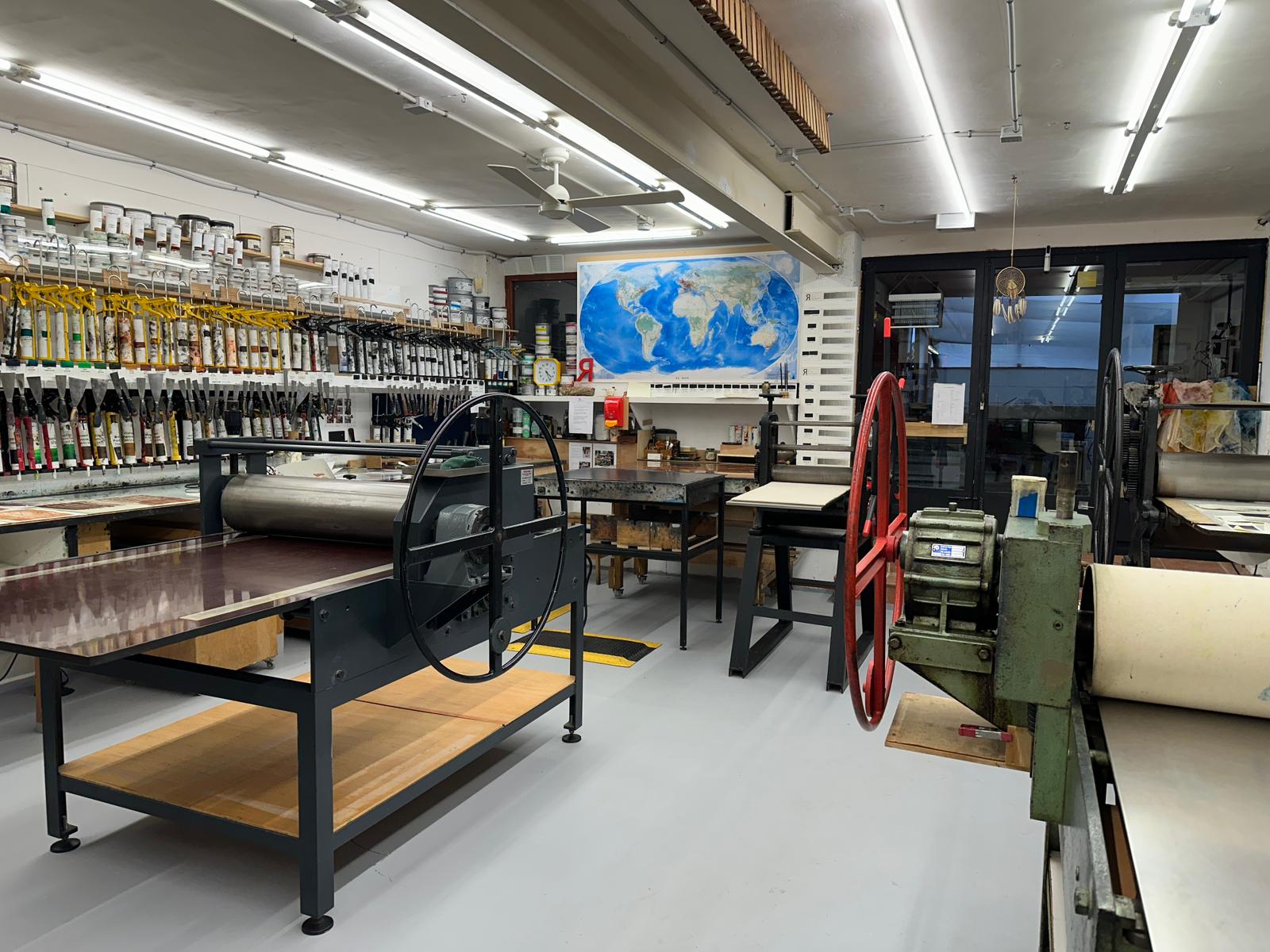 A group of people gathered around the etching press at Hello Print Friend Studio surrounded by large windows letting in lots of natural light.