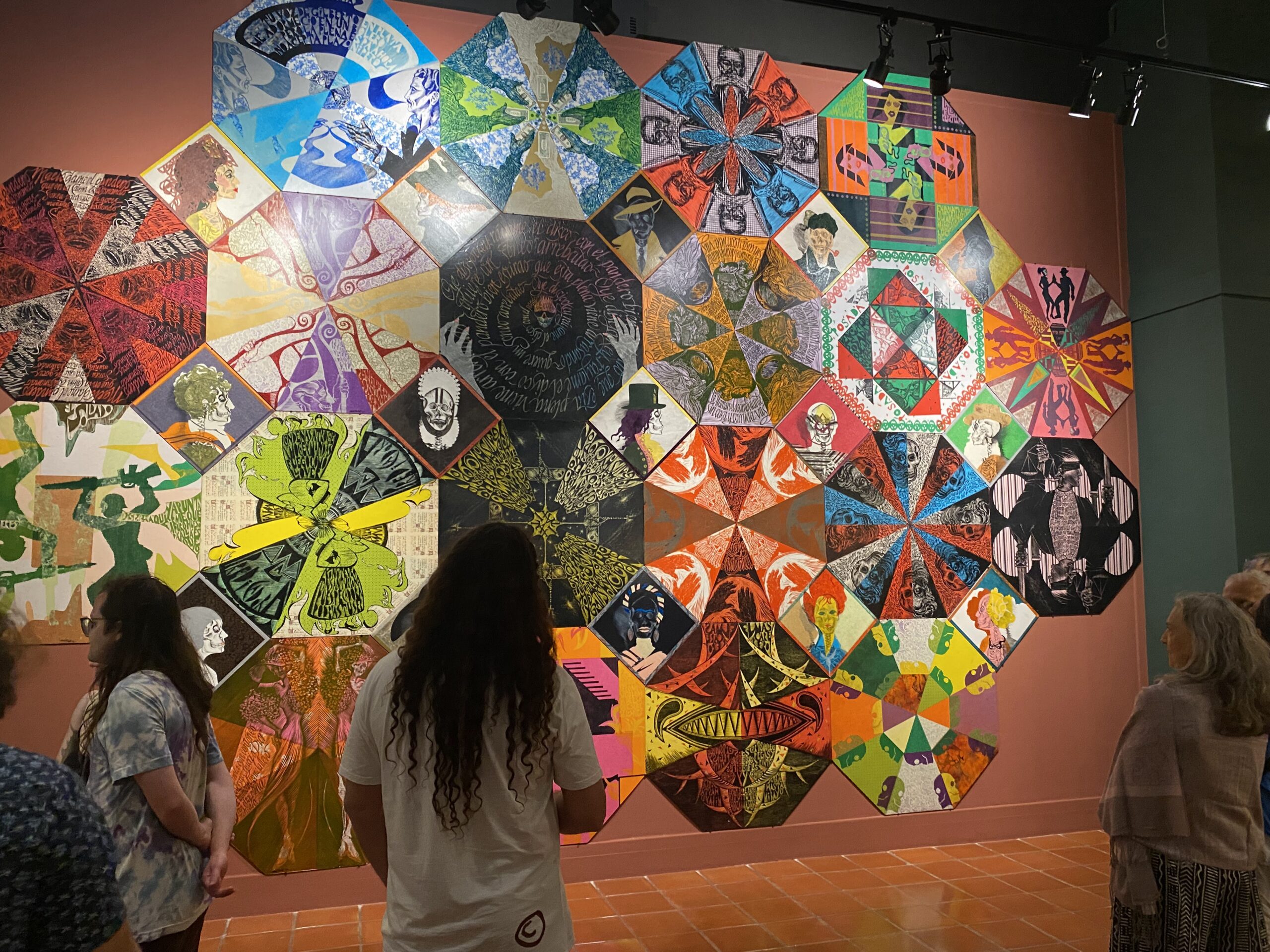 Visitors view a large collaborative installation of relief-printed artworks, arranged as overlapping geometric forms across a pink gallery wall. The prints feature vivid colors, portraits, patterns, text, and radial compositions.